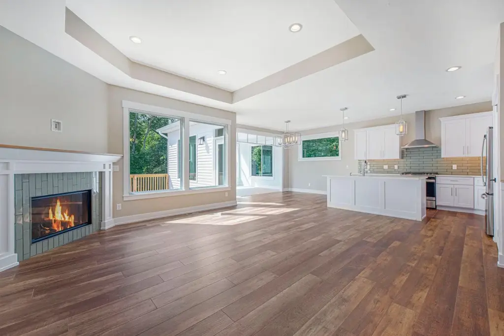 Open-concept custom home living room and kitchen with a fireplace, large windows, white cabinetry, and wood flooring