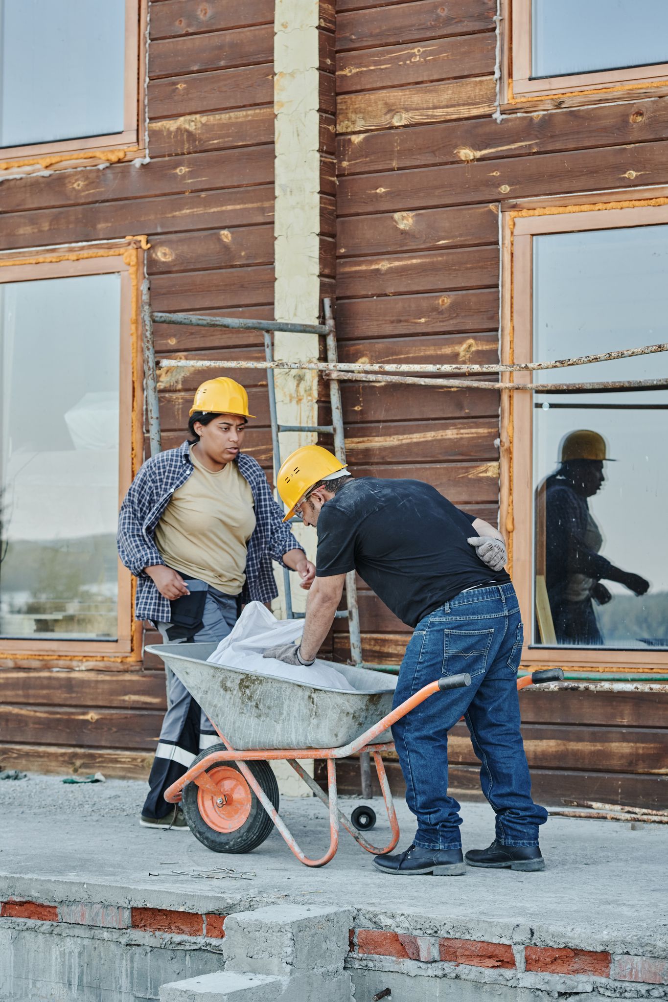 Two construction workers in hard hats at a wooden house, inspecting work plans in a wheelbarrow.