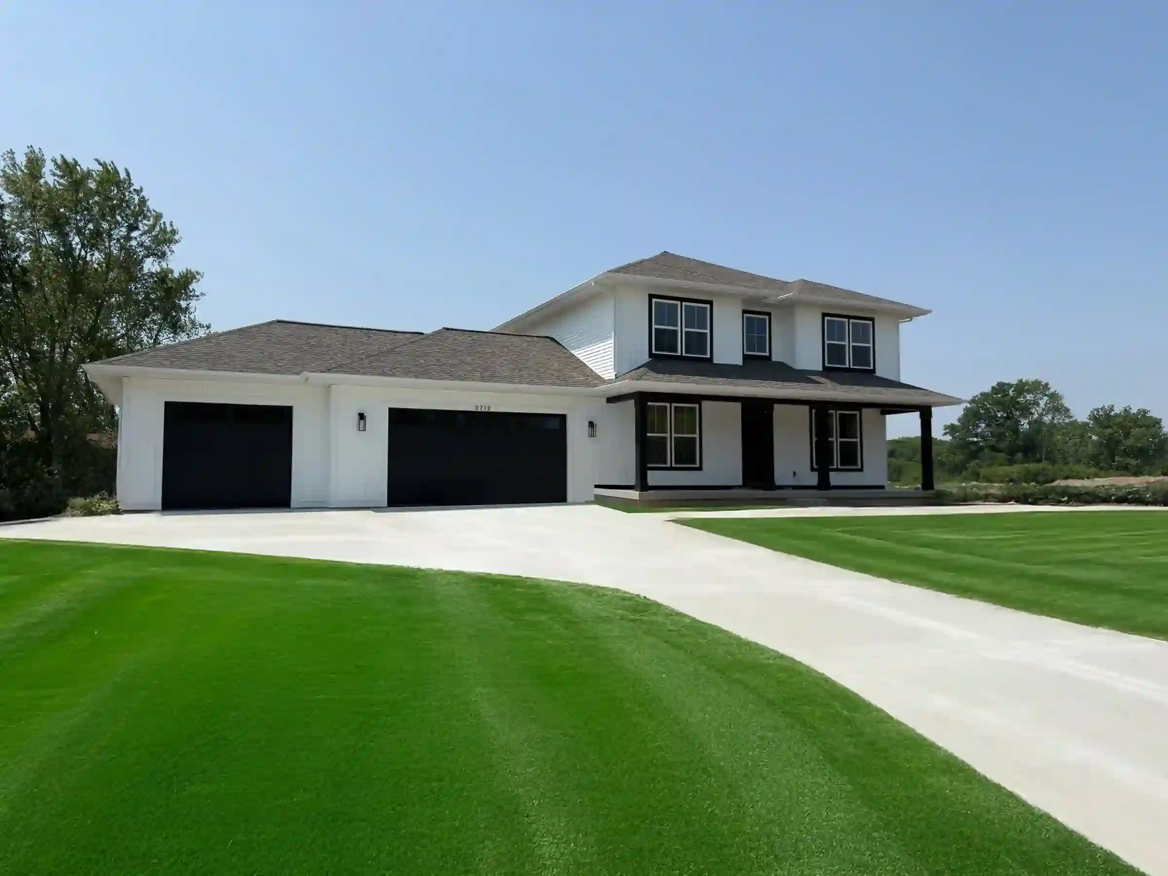 White modern farmhouse custom home with black garage doors, covered porch, curved driveway, and green lawn in West Michigan