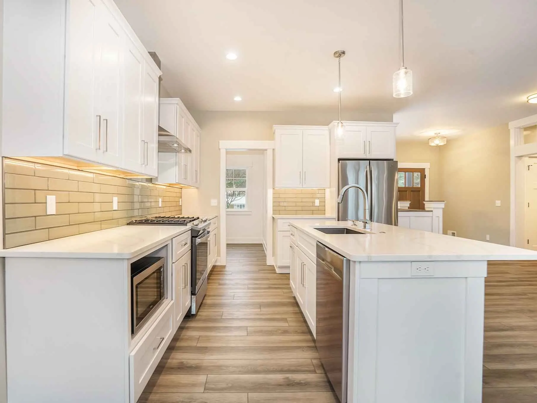 Bright custom kitchen with white shaker cabinets, quartz island, stainless steel appliances, subway tile backsplash, and wood-look flooring