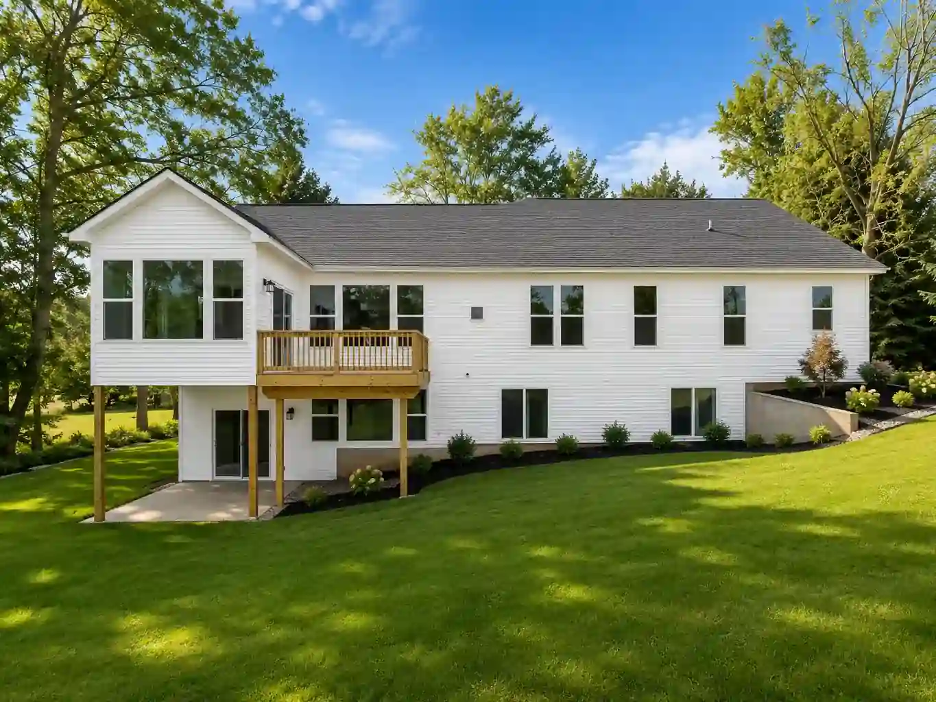 Rear exterior of a white custom home with an elevated wood deck, walkout patio, landscaped slope, and green lawn in West Michigan