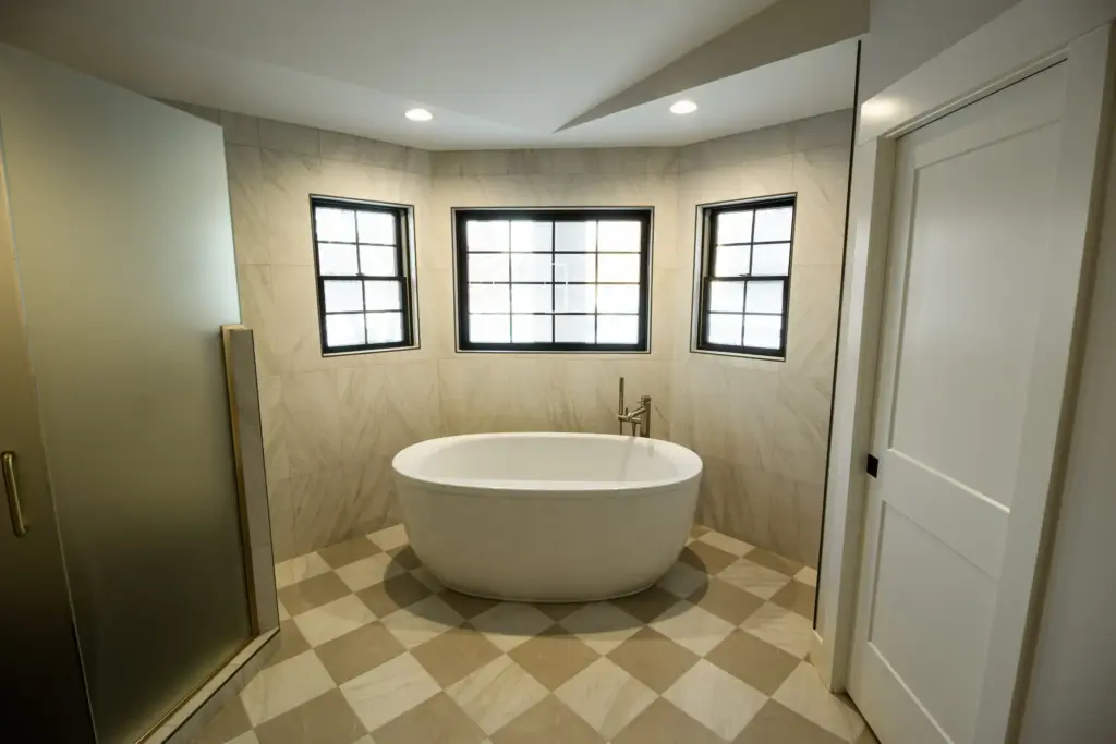 Freestanding soaking tub beneath three black-trimmed windows in remodeled bathroom