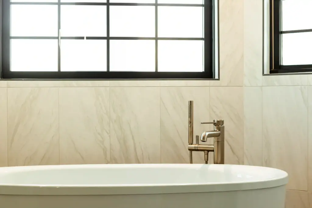 Freestanding tub with brushed metal faucet beneath black-trimmed bathroom windows