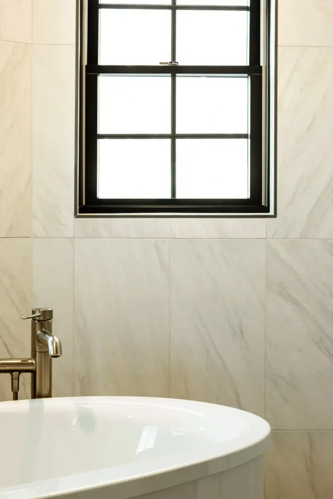 Freestanding tub beneath a black-trimmed window and marble-look wall tile